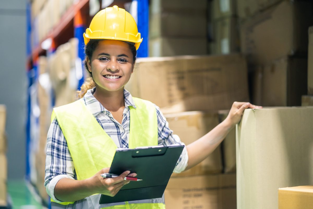 Young woman worker in safety helmet and vest checking box in stock in warehouse.